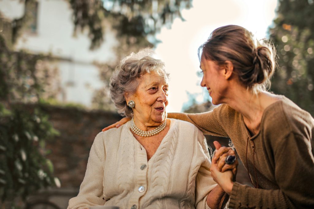 Mulher cuida de idosa com carinho. Elas sorriem em um ambiente aberto, cercado de verde.