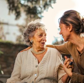Mulher cuida de idosa em ambiente aberto cercado de verde. As duas sorriem